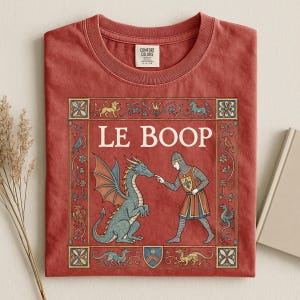 May include: A brick red t-shirt featuring a medieval-style graphic with the text LE BOOP. The illustration depicts a knight in chainmail touching the nose of a blue dragon, framed by a decorative border with small mythical creatures and floral motifs.