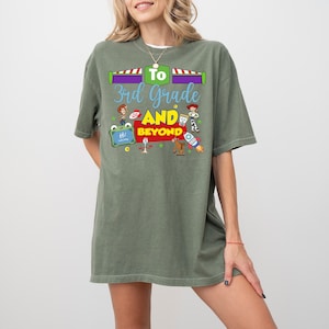 May include: An olive green short-sleeve t-shirt featuring a graphic print with the text "To 3rd Grade AND BEYOND". The design includes colorful illustrations of various cartoon characters, a rocket ship, and a small chalkboard with the word "Hi".