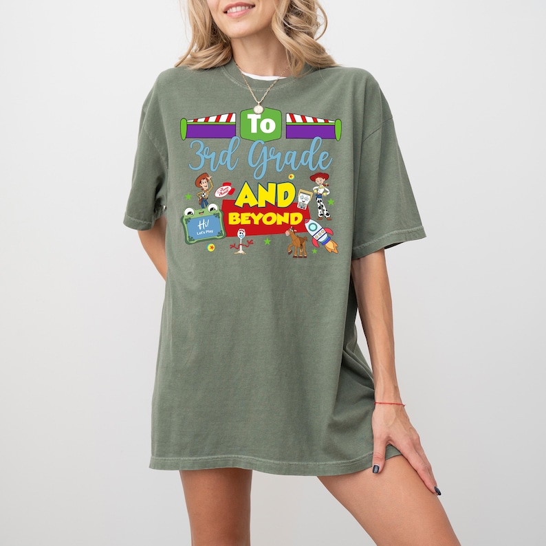 May include: An olive green short-sleeve t-shirt featuring a graphic print with the text "To 3rd Grade AND BEYOND". The design includes colorful illustrations of various cartoon characters, a rocket ship, and a small chalkboard with the word "Hi".