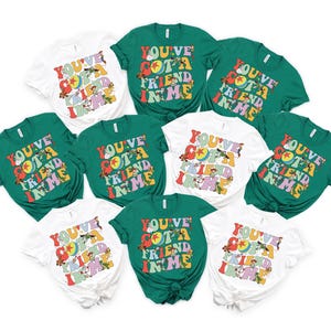May include: A collection of white and green t-shirts with the text "YOU'VE GOT A FRIEND IN ME" in a retro font. The shirts feature cartoon characters and a colorful design, perfect for fans of the movie.