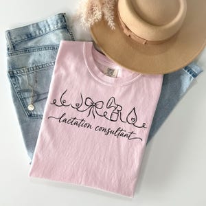 May include: A light pink t-shirt featuring a black line art graphic of breasts, a bow, a breast pump, and a water drop, with the text "lactation consultant" written in cursive script below the icons. The shirt is paired with light blue denim jeans.