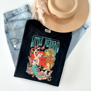 May include: Black t-shirt featuring a colorful graphic of Ariel and Flounder from The Little Mermaid, with the text "Little Mermaid". The shirt is paired with light wash jeans and a tan hat.