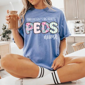 以下が含まれることがあります： 「TINY PATIENTS, BIG HEARTS」と「PEDS nurse」の文字がカラフルな花柄でデザインされた、水色のTシャツ。人物はストローと茶色の液体が入ったガラス瓶を持っています。