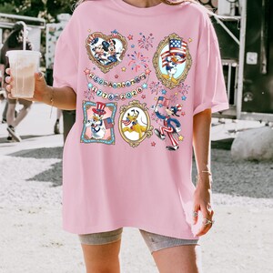 Pode incluir: T-shirt rosa claro com personagens de desenhos animados em trajes patri&oacute;ticos. O design inclui imagens emolduradas de Mickey Mouse, Pato Donald e Pluto, e o texto "250th Anniversary 1776-2026".