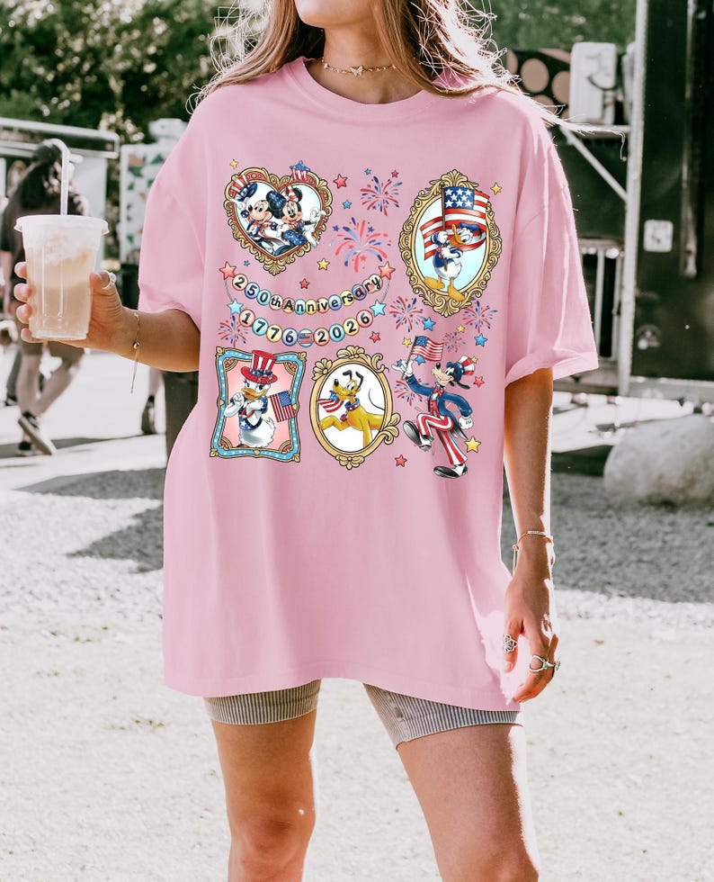 Pode incluir: T-shirt rosa claro com personagens de desenhos animados em trajes patri&oacute;ticos. O design inclui imagens emolduradas de Mickey Mouse, Pato Donald e Pluto, e o texto "250th Anniversary 1776-2026".