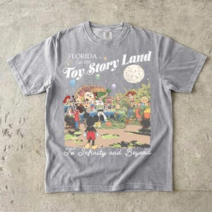 May include: A grey short-sleeve t-shirt featuring a graphic print of Toy Story Land with characters including Mickey Mouse, Woody, and Buzz Lightyear. The text reads Florida Est 2018, Toy Story Land, and To Infinity and Beyond in white script.