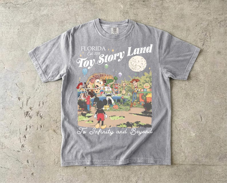 May include: A grey short-sleeve t-shirt featuring a graphic print of Toy Story Land with characters including Mickey Mouse, Woody, and Buzz Lightyear. The text reads Florida Est 2018, Toy Story Land, and To Infinity and Beyond in white script.
