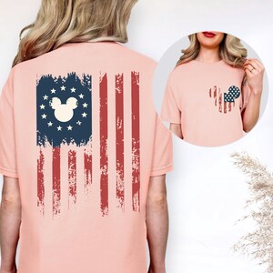 Puede incluir: Una camiseta color melocot&oacute;n con un gr&aacute;fico de la bandera estadounidense desgastada en la espalda, que incluye una silueta de orejas de rat&oacute;n rodeada de estrellas. El pecho muestra una silueta de orejas de rat&oacute;n m&aacute;s peque&ntilde;a y a juego.