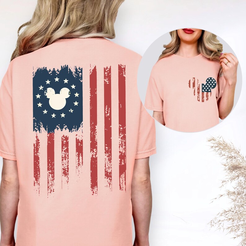 Puede incluir: Una camiseta color melocot&oacute;n con un gr&aacute;fico de la bandera estadounidense desgastada en la espalda, que incluye una silueta de orejas de rat&oacute;n rodeada de estrellas. El pecho muestra una silueta de orejas de rat&oacute;n m&aacute;s peque&ntilde;a y a juego.