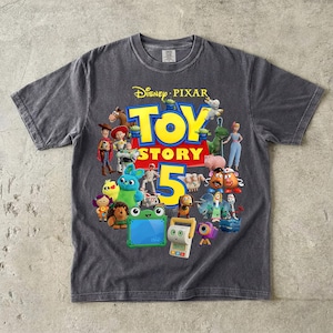 May include: A charcoal gray short-sleeve t-shirt featuring a graphic print of various Toy Story characters surrounding the large yellow text Toy Story 5 and the Disney Pixar logo at the top. The shirt has a crew neckline and a vintage-style wash.