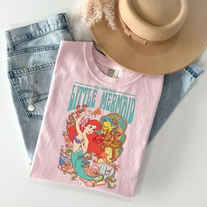 May include: A light pink t-shirt featuring a graphic of Ariel and Flounder from The Little Mermaid, with the words "Little Mermaid" in teal. The shirt is paired with light wash jeans, a tan hat, and a necklace.