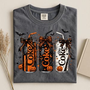 Puede incluir: Una camiseta gris carbón con un gráfico de tres latas de Diet Coke decoradas para Halloween. Cada lata tiene una pajita, un lazo y patrones de telaraña. Las latas son de color naranja, negro y blanco, rodeadas de calabazas y murciélagos.