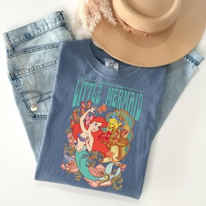 May include: A blue t-shirt featuring a graphic of Ariel and Flounder from The Little Mermaid. The shirt is laid on top of a pair of light-wash jeans, a gold necklace, and a tan hat. The shirt has the words "Little Mermaid" printed on it.