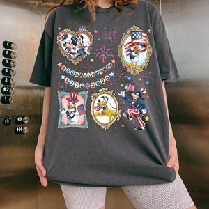 Pode incluir: T-shirt cinzenta escura com personagens de desenhos animados em molduras patri&oacute;ticas, fogos de artif&iacute;cio e o texto "250th Anniversary 1776-2026". A camisa tem um ajuste folgado e mangas curtas. O design inclui Mickey e Minnie Mouse, Pato Donald e Pluto.