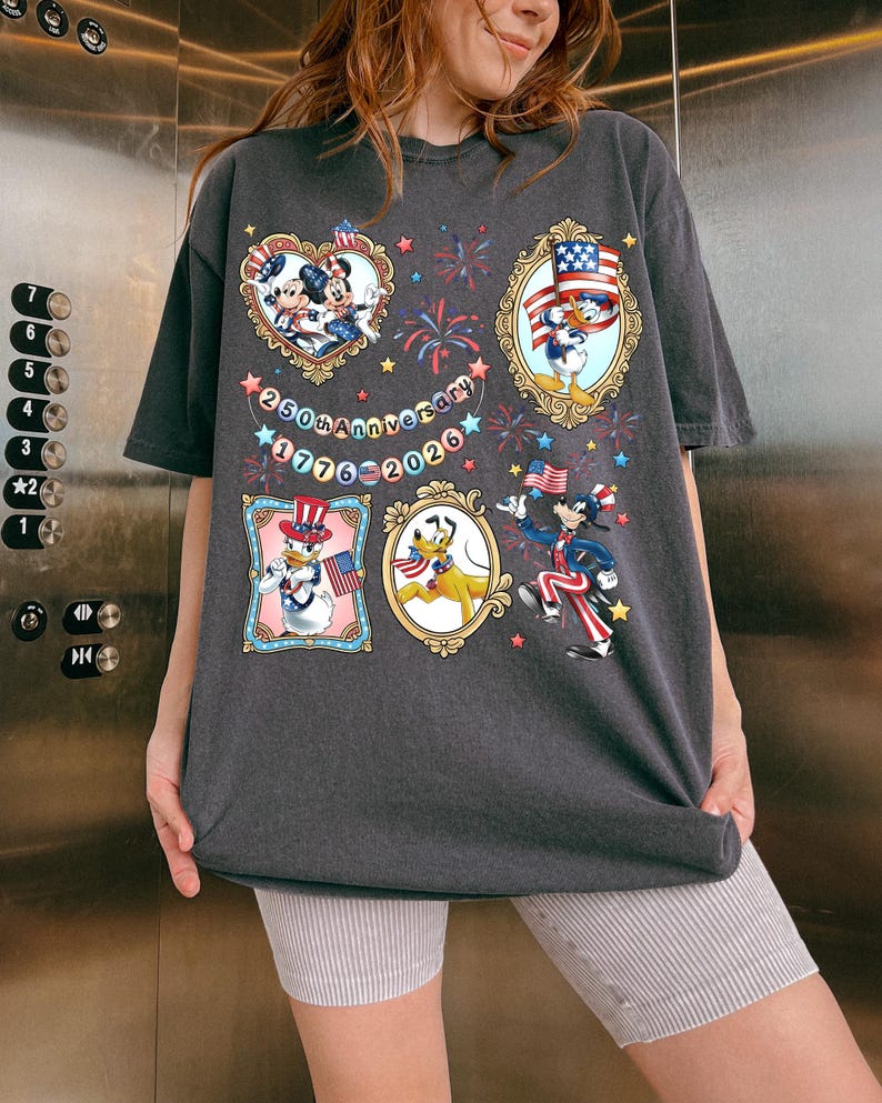 Pode incluir: T-shirt cinzenta escura com personagens de desenhos animados em molduras patri&oacute;ticas, fogos de artif&iacute;cio e o texto "250th Anniversary 1776-2026". A camisa tem um ajuste folgado e mangas curtas. O design inclui Mickey e Minnie Mouse, Pato Donald e Pluto.