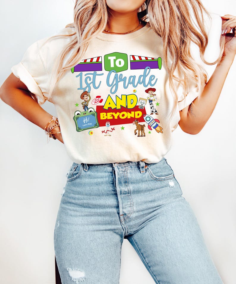 May include: A cream-colored t-shirt featuring a colorful graphic with the text To 1st Grade AND BEYOND. The design includes illustrations of various toy characters, a rocket ship, and a green frame with the text Hi! Let's Play, set against a light background.