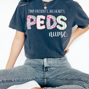 以下が含まれることがあります： 「TINY PATIENTS. BIG HEARTS」の文字がプリントされた濃い青色のTシャツ。 「PEDS」の文字は花柄とチェック柄のデザインで、その下に「nurse」の文字が配置されています。 青いジーンズと白い靴と合わせています。