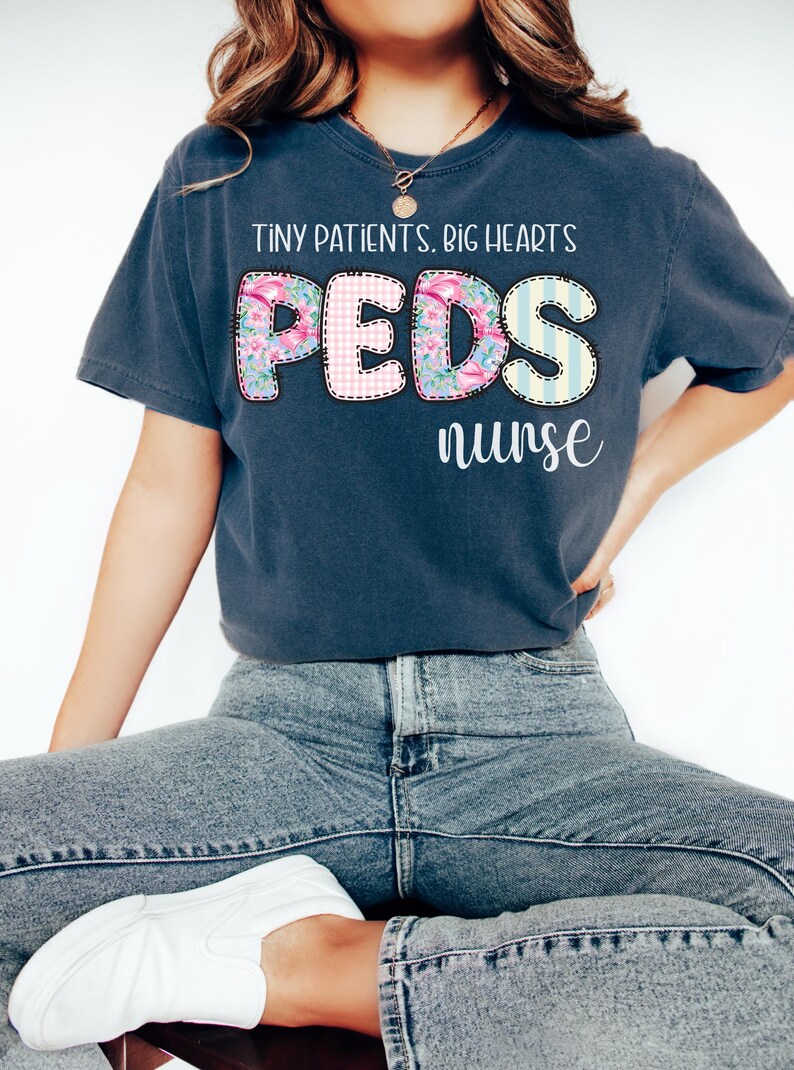 以下が含まれることがあります： 「TINY PATIENTS. BIG HEARTS」の文字がプリントされた濃い青色のTシャツ。 「PEDS」の文字は花柄とチェック柄のデザインで、その下に「nurse」の文字が配置されています。 青いジーンズと白い靴と合わせています。