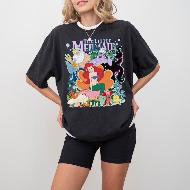 May include: Black t-shirt featuring a colorful graphic of characters from "The Little Mermaid." The design includes Ariel, King Triton, Ursula, and other characters. The shirt has a white crew neck collar and short sleeves.