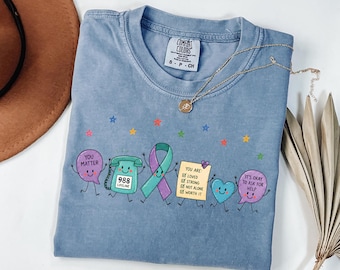 May include: A blue crew neck t-shirt featuring a graphic print of cartoon characters including a speech bubble with the text YOU MATTER, a telephone with 988 LIFELINE, a ribbon, a checklist with YOU ARE: LOVED, STRONG, NOT ALONE, WORTH IT, and a heart.