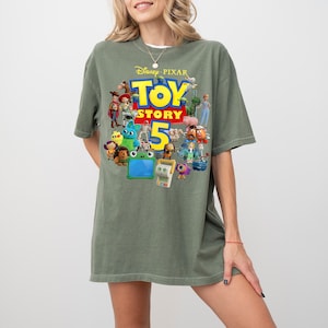 May include: An olive green short-sleeve t-shirt featuring a graphic print of various Toy Story characters surrounding the yellow and red text Toy Story 5. The design includes figures like Woody, Buzz Lightyear, Jessie, and others in a collage.