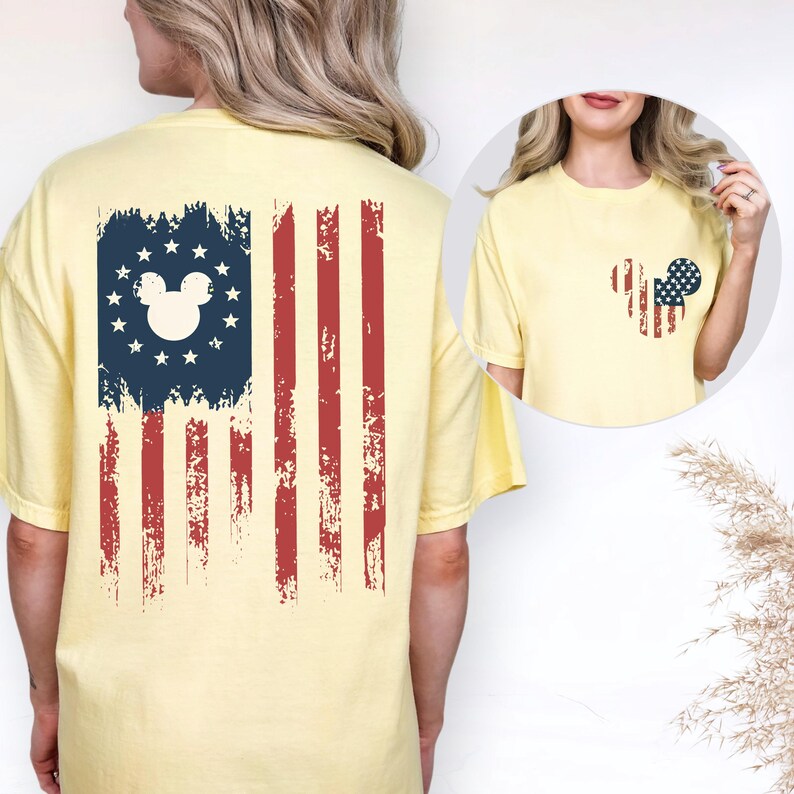 Puede incluir: Una camiseta de manga corta de color amarillo p&aacute;lido con un gr&aacute;fico de la bandera estadounidense desgastada en la espalda, con una silueta central de Mickey Mouse rodeada de estrellas. El pecho muestra una silueta de Mickey Mouse m&aacute;s peque&ntilde;a.