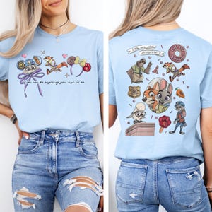 May include: A light blue t-shirt featuring Zootopia character graphics. The front displays ears, a fox, and the text 'You can be anything you wish to be.' The back shows a collage of characters, a badge, a donut, and a carrot graphic.
