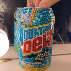 May include: A bedazzled Mountain Dew Baja Blast can. The can is covered in iridescent rhinestones, with the Mountain Dew logo in red and white. The can also has the words "Baja Blast Tropical Lime" on it.