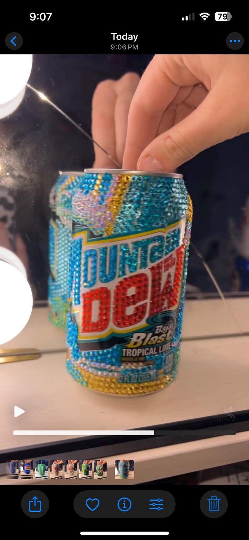 May include: A bedazzled Mountain Dew Baja Blast can. The can is covered in iridescent rhinestones, with the Mountain Dew logo in red and white. The can also has the words "Baja Blast Tropical Lime" on it.