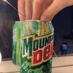 May include: A bedazzled Mountain Dew can, reflecting in a mirror. The can is covered in green, gold, and iridescent rhinestones. The Mountain Dew logo is visible in green and red. The can is held by a hand.
