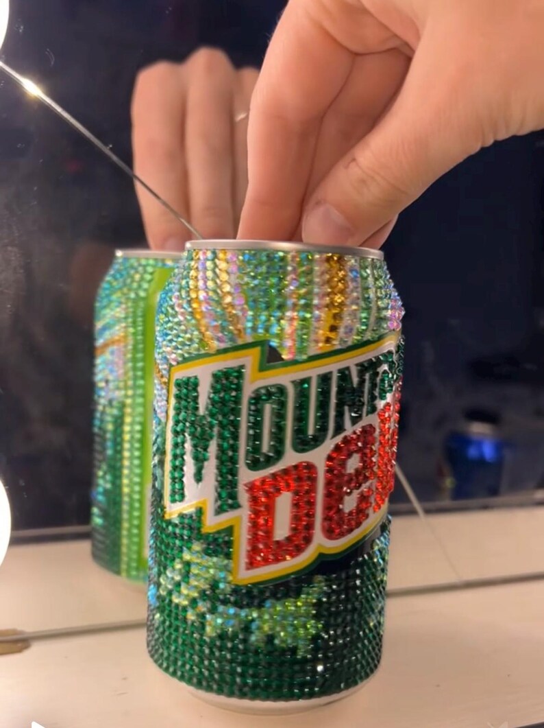 May include: A bedazzled Mountain Dew can, reflecting in a mirror. The can is covered in green, gold, and iridescent rhinestones. The Mountain Dew logo is visible in green and red. The can is held by a hand.