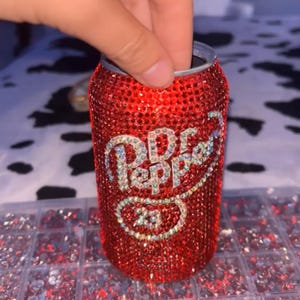 May include: A red Dr. Pepper can covered in sparkling rhinestones. The Dr. Pepper logo is visible in the center of the can. The can is held by a hand. Rhinestones are in a clear plastic container.