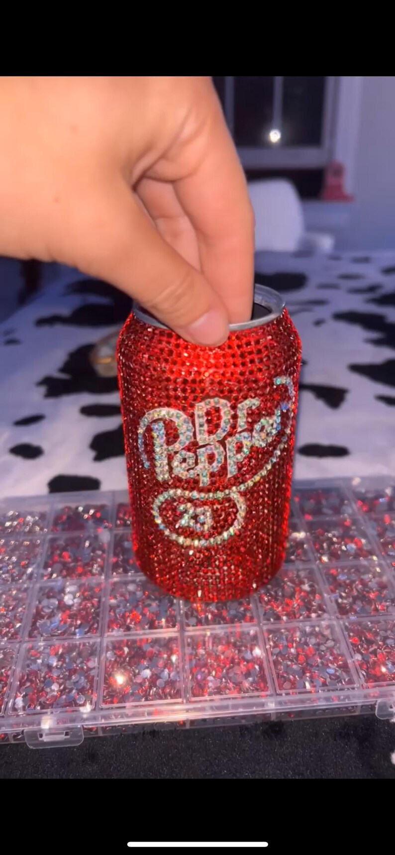 May include: A red Dr. Pepper can covered in sparkling rhinestones. The Dr. Pepper logo is visible in the center of the can. The can is held by a hand. Rhinestones are in a clear plastic container.