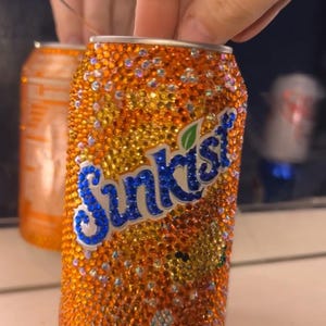 May include: Orange Sunkist soda can covered in orange and clear rhinestones. The Sunkist logo is in blue with a green leaf. A second, similar can is visible in the background.