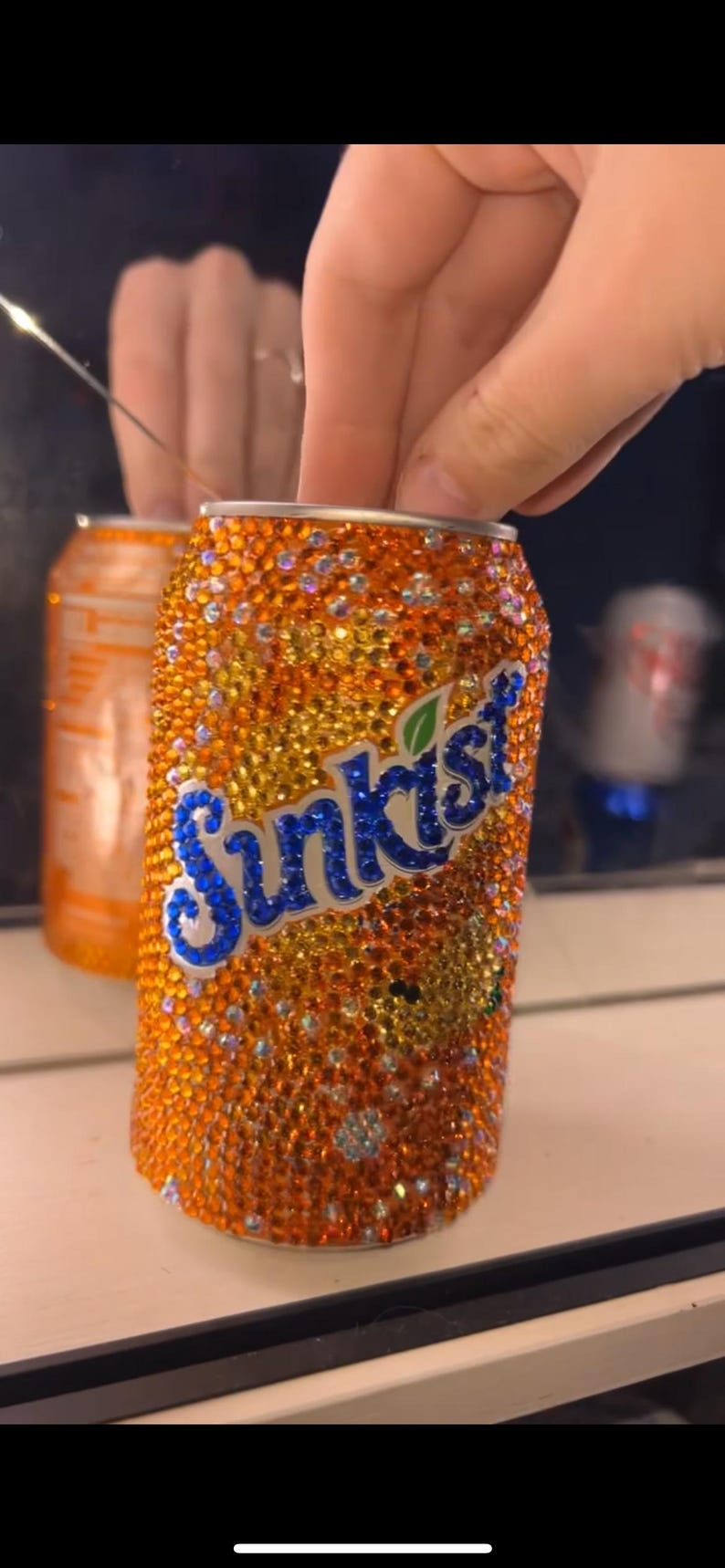 May include: Orange Sunkist soda can covered in orange and clear rhinestones. The Sunkist logo is in blue with a green leaf. A second, similar can is visible in the background.