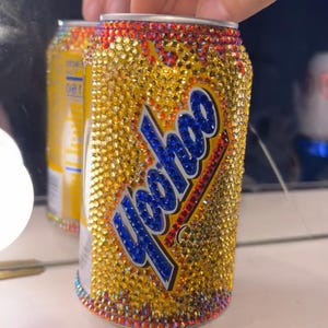Puede incluir: Una lata de Yoohoo adornada con pedrería dorada. El texto "Yoohoo" es azul con un contorno blanco. Los bordes superior e inferior son rojos. La lata se sostiene frente a un espejo.