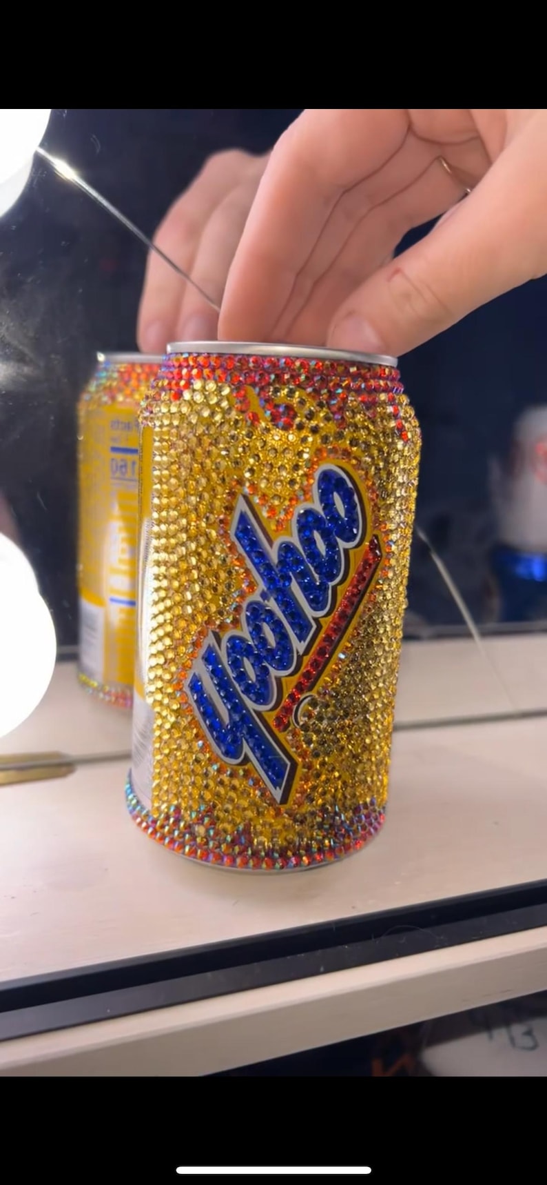 May include: A bedazzled can of Yoohoo, covered in gold rhinestones. The text "Yoohoo" is in blue with a white outline. The top and bottom rims are red and the can is held in front of a mirror.