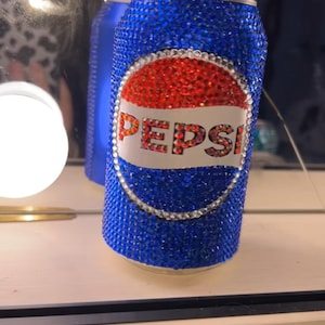 May include: A bedazzled Pepsi can, covered in blue rhinestones, with a red and white Pepsi logo. The can is held by a person. The can is a unique, decorative item.
