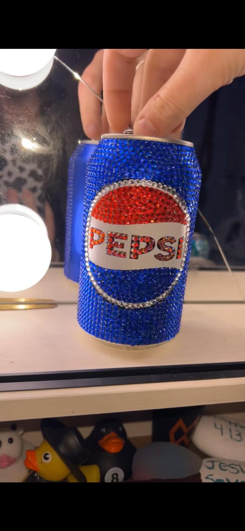 May include: A bedazzled Pepsi can, covered in blue rhinestones, with a red and white Pepsi logo. The can is held by a person. The can is a unique, decorative item.