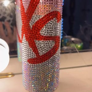 May include: A bedazzled beverage can with a red "KS" design. The can is covered in iridescent rhinestones, with the top reading "VITAMIN C". The design is eye-catching and decorative.