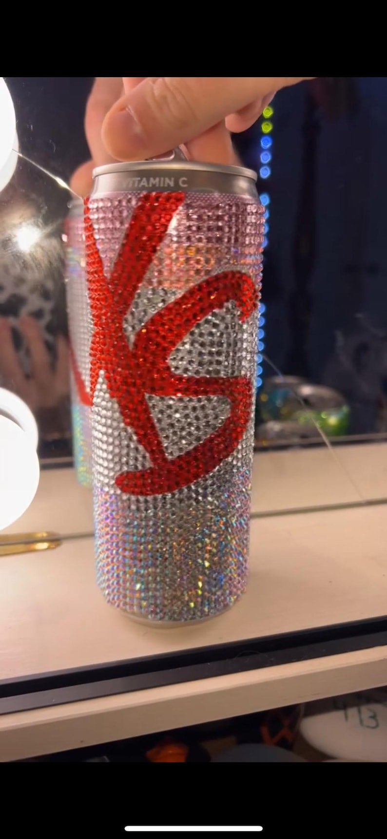 May include: A bedazzled beverage can with a red "KS" design. The can is covered in iridescent rhinestones, with the top reading "VITAMIN C". The design is eye-catching and decorative.