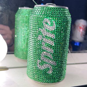 May include: A green soda can covered in sparkling green rhinestones. The word "Sprite" is spelled out in silver rhinestones on the front of the can. The can is reflected in a mirror.