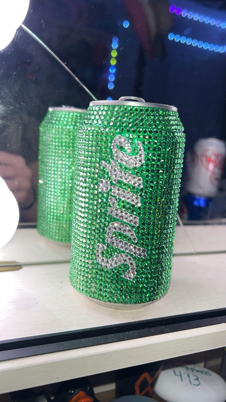 May include: A green soda can covered in sparkling green rhinestones. The word "Sprite" is spelled out in silver rhinestones on the front of the can. The can is reflected in a mirror.