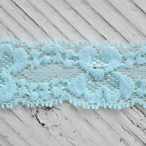 May include: A close-up of a light blue lace trim with a delicate floral pattern. The lace is on a white wooden surface.