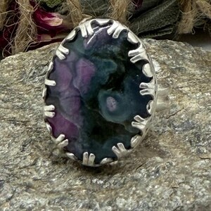 Puede incluir: Un anillo de plata con una piedra ovalada que presenta un patrón jaspeado en tonos púrpura intenso, verde oscuro y negro. La piedra está asegurada por un engaste de plata decorativo con pequeños acentos metálicos dentados en el borde.