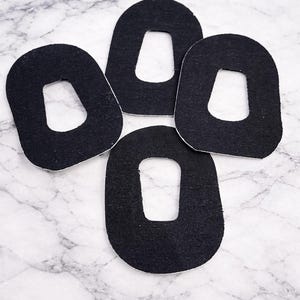 May include: Four black, oval-shaped felt pads with a white border are arranged on a marble surface. Each pad has a rectangular cutout in the center. The pads are evenly spaced and slightly angled.