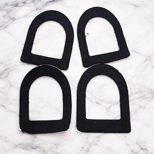 Può includere: Quattro forme in feltro nero, ciascuna con la parte superiore arrotondata e la parte inferiore piatta, disposte su una superficie di marmo bianco e grigio. Le forme sono identiche, simili a cornici ad arco o elementi decorativi.