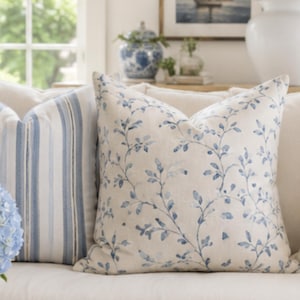 Könnte beinhalten: Zwei quadratische Dekokissen auf einem beigen Sofa. Ein Kissen hat vertikale blaue und weiße Streifen, während das andere ein blaues botanisches Rankenmuster auf cremefarbenem Grund zeigt. Die Kissen stehen neben einer blau-weißen Blumenvase.