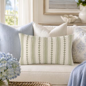 May include: A rectangular throw pillow featuring a cream background with vertical sage green stripes and a repeating leaf pattern. It is positioned on a sofa alongside a solid light blue square pillow and a white square pillow with a floral print.