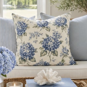 Könnte beinhalten: Ein quadratisches Zierkissen mit einem blauen Hortensien-Blumenmuster auf cremefarbenem Hintergrund. Das Kissen wird neben einem blau-weiß gestreiften Kissen und einem einfarbigen hellblauen Kissen auf einem geflochtenen Möbelstück präsentiert.